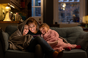 Two young girls with their mother, sitting comfortably with a blanket over their heads on a sofa in the living room of their home